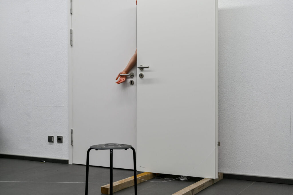 A partially open white door with a hand reaching out, beside a black stool on a wooden base, in a minimalist room.