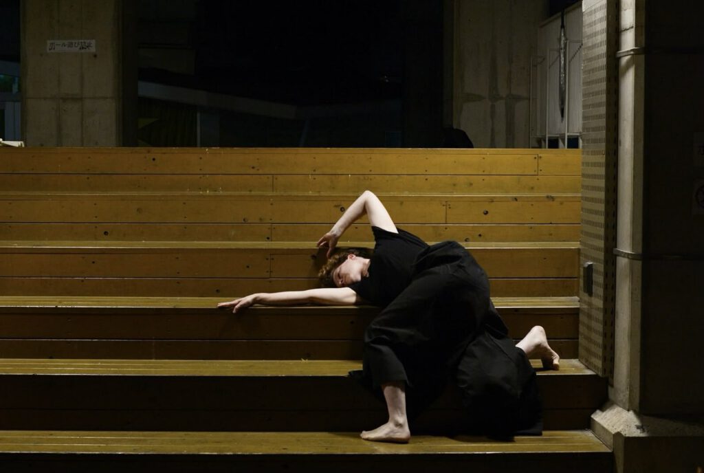 A dancer in black clothing performs on wooden steps, arching gracefully with one arm extended, in a dimly lit setting.