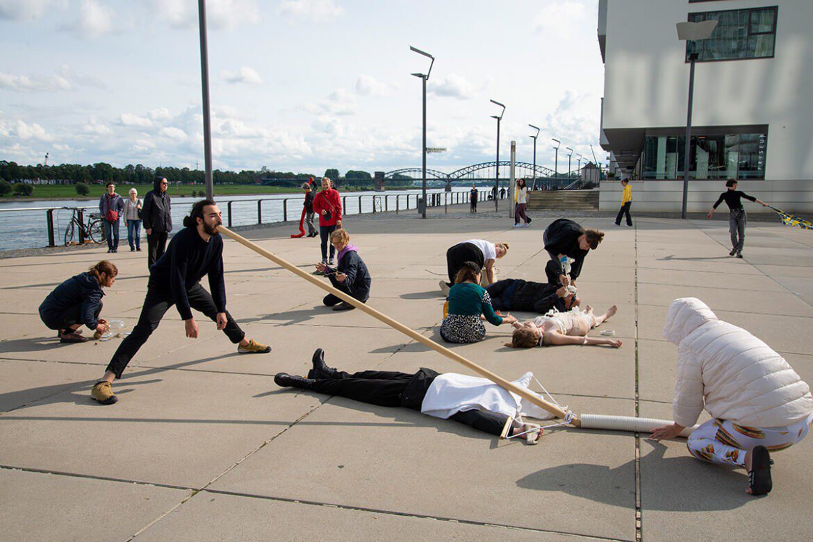 People participating in a dramatic performance on a riverside walkway, some lying on the ground and others interacting with props.