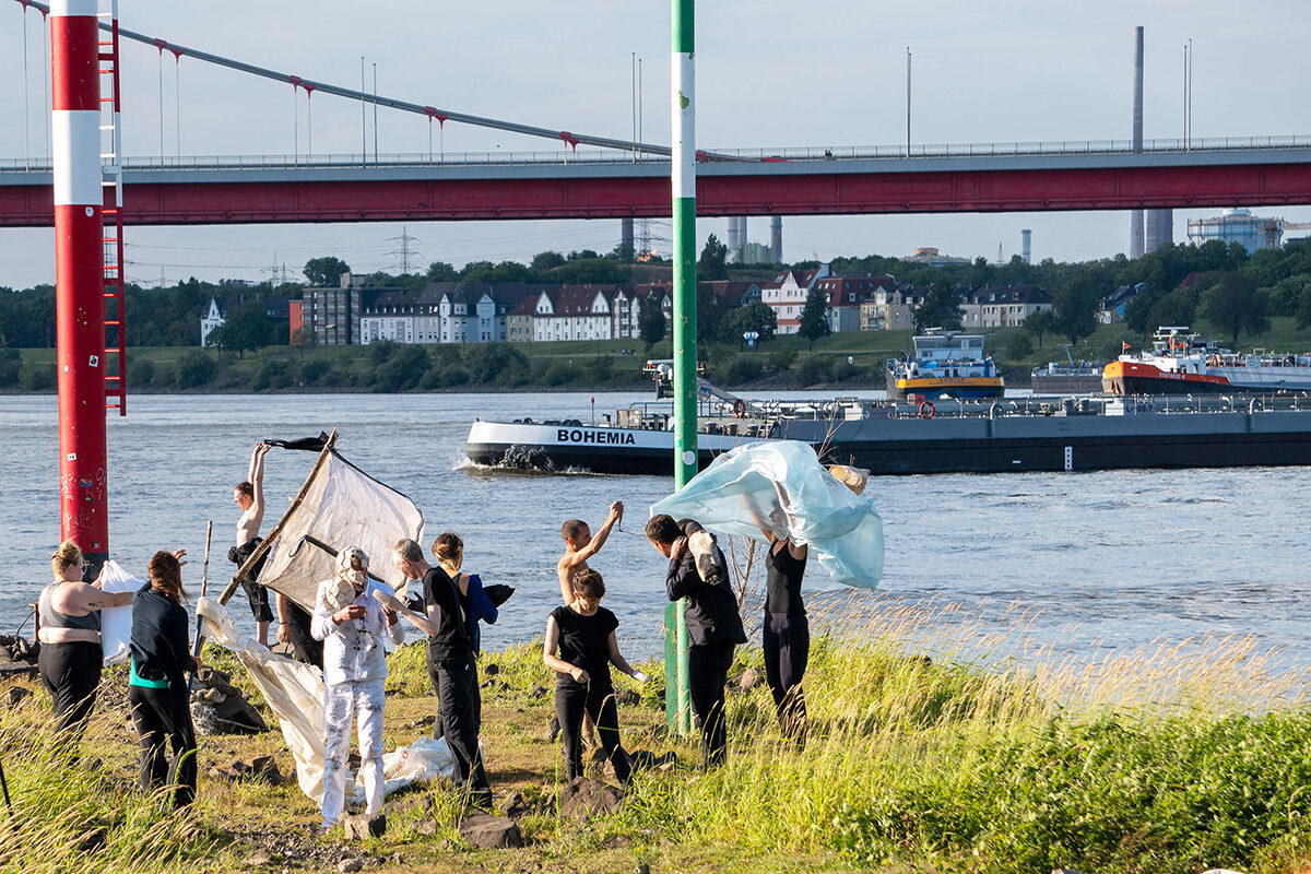 People in black clothing hold up large fabrics near a river, with a barge passing under a bridge in the background.