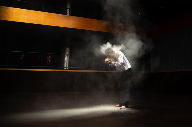 A performer in a white shirt bows in a spotlight, surrounded by a cloud of smoke, conveying a dramatic, introspective mood.