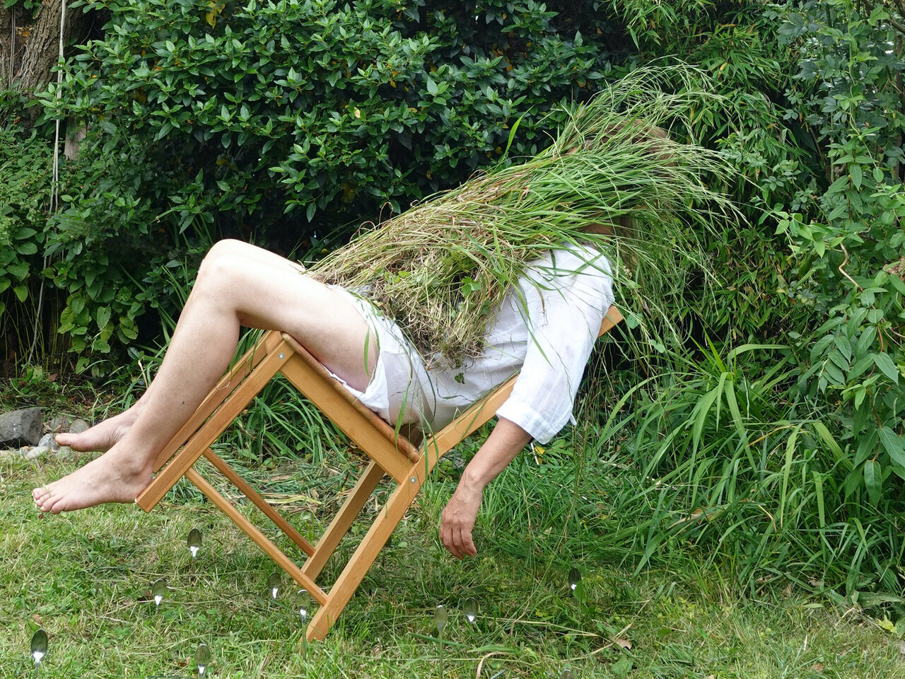 Person reclines in a chair with grass obscuring their head, surrounded by lush greenery, creating a surreal and humorous scene.