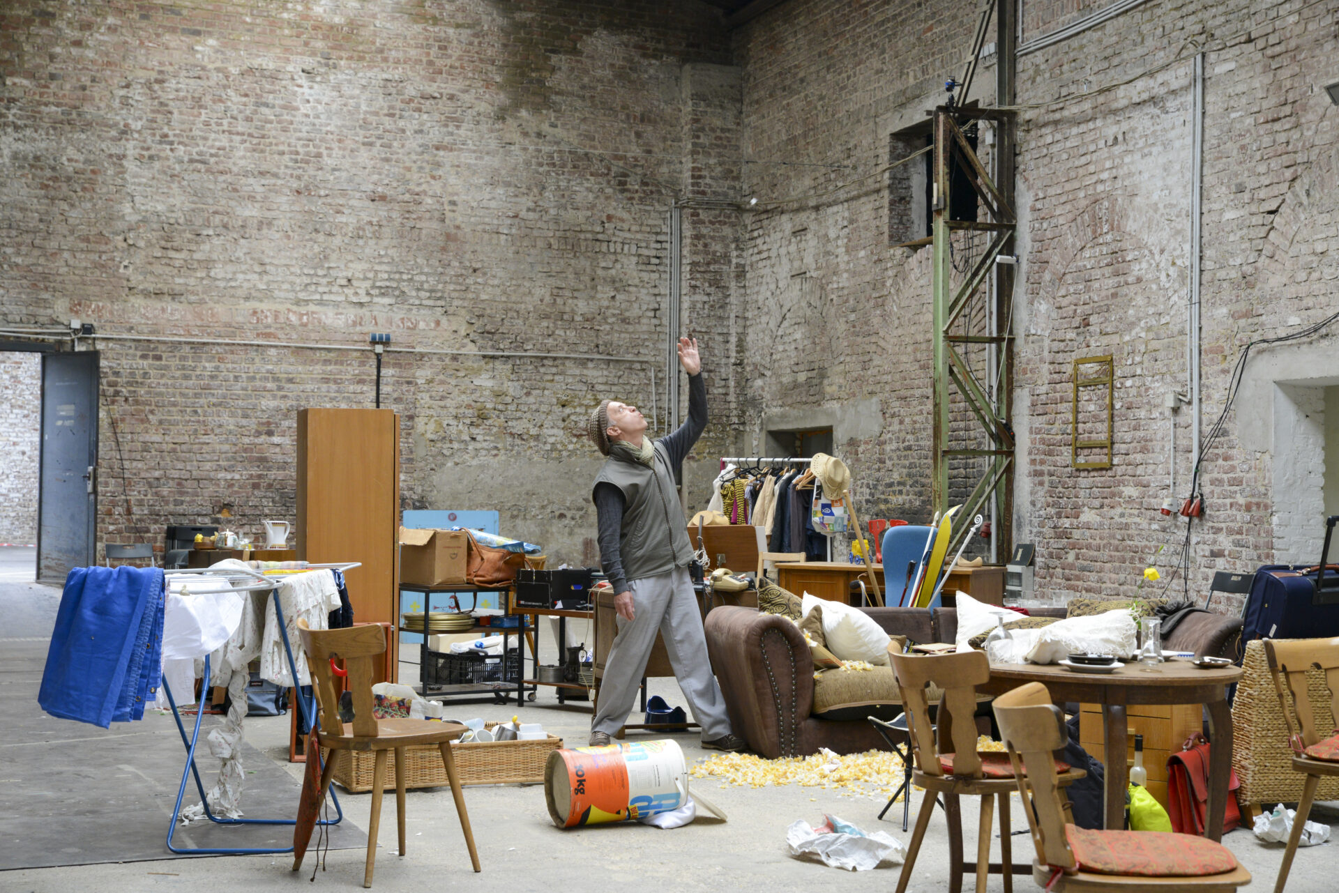 A person gestures upward in a cluttered, industrial space filled with furniture and discarded items, amid exposed brick walls.