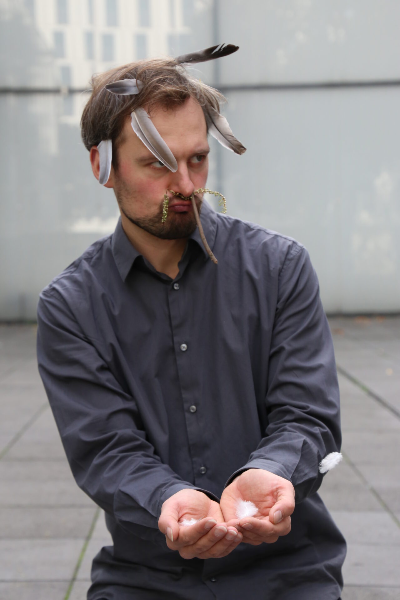 Man with a serious expression, wearing a gray shirt, holds his hands out as feathers and strips of paper float around him.