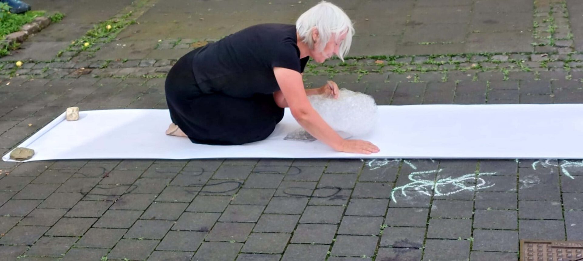 A person kneels on a white sheet outdoors, drawing with chalk while surrounded by stone pavement and greenery.