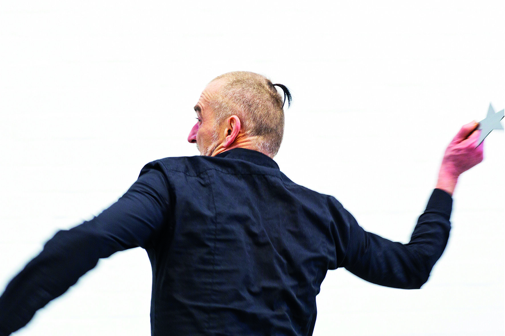 A man with a shaved head and ponytail throws a star-shaped object against a plain white wall.