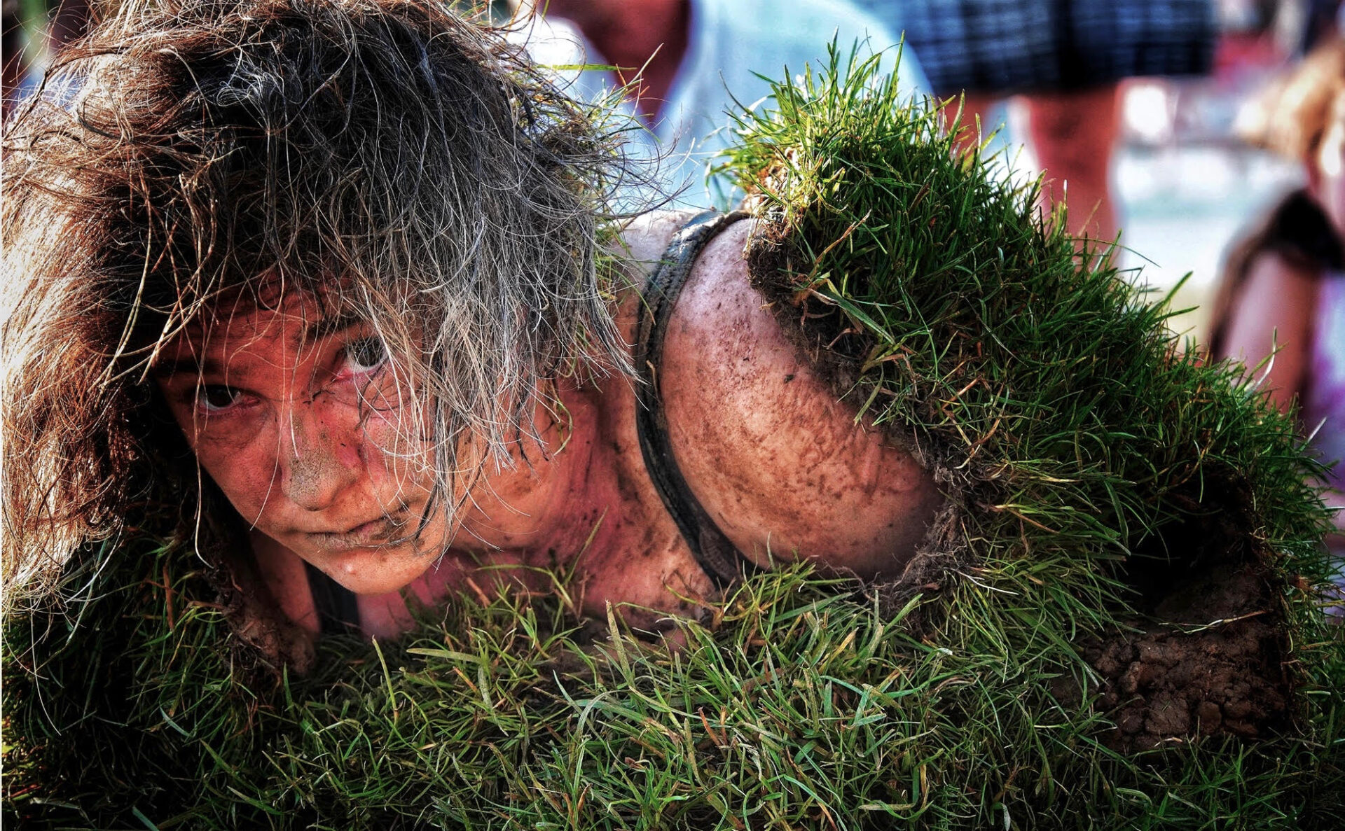 Person covered in grass and dirt, emerging from the ground, with intense expression; spectators in background.