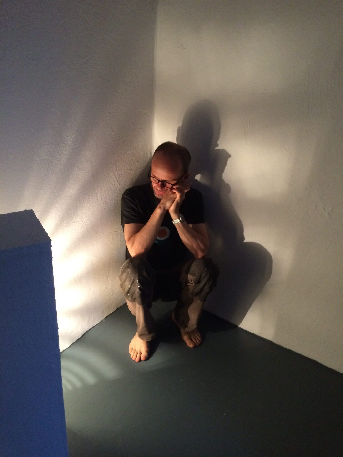 A person sitting in a corner, holding their head with hands, barefoot on a blue floor, casting long shadows against the wall.