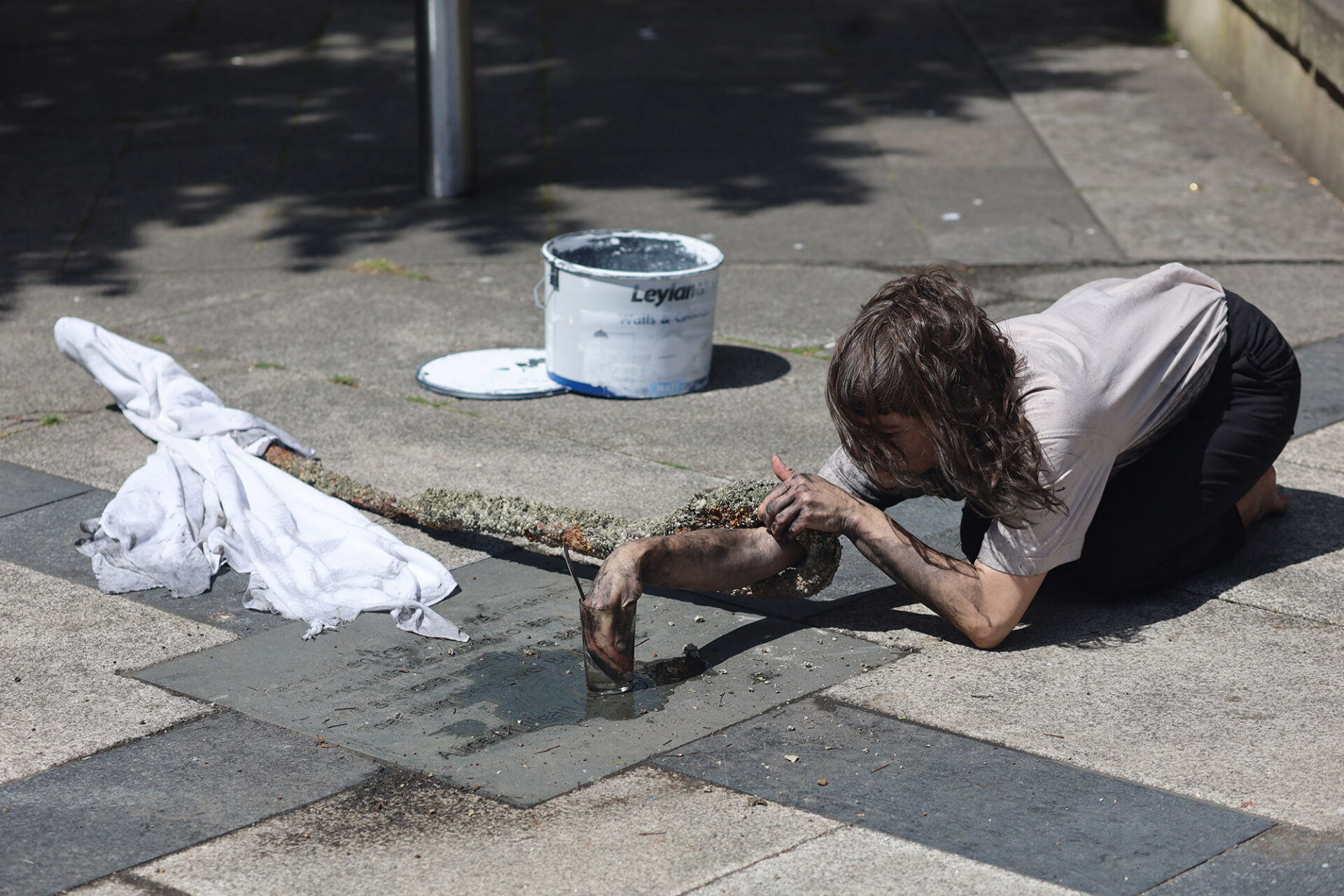 A person in a t-shirt kneels on a pavement, scrubbing a concrete surface with a cloth, a paint bucket nearby.