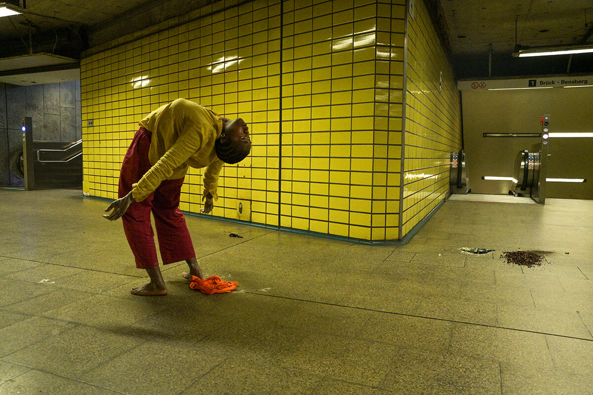A person in a yellow sweater and red pants bends down near a bright yellow tiled wall, with a subway entrance in the background.