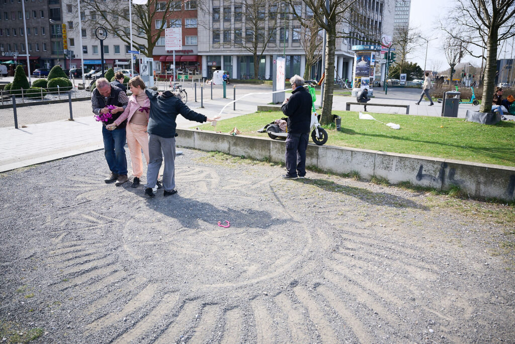 Four people stand around a large sand design in a public park, with trees and buildings in the background.