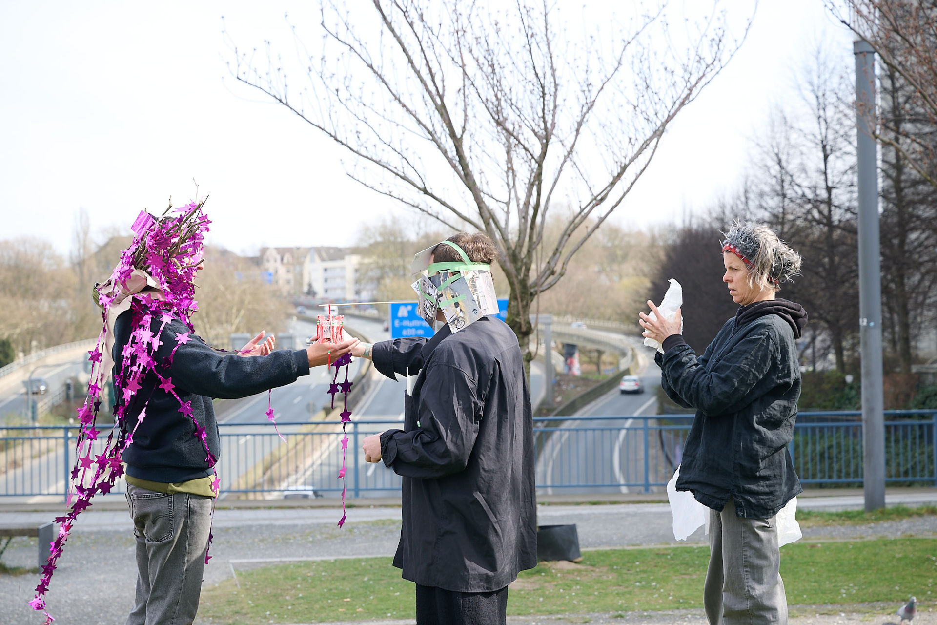 Zwei Personen sind in eine Outdoor-Performance involviert, wobei eine eine aus Fotos gefertigte Maske trägt und eine Frucht hält, während die andere in der Nähe interagiert.