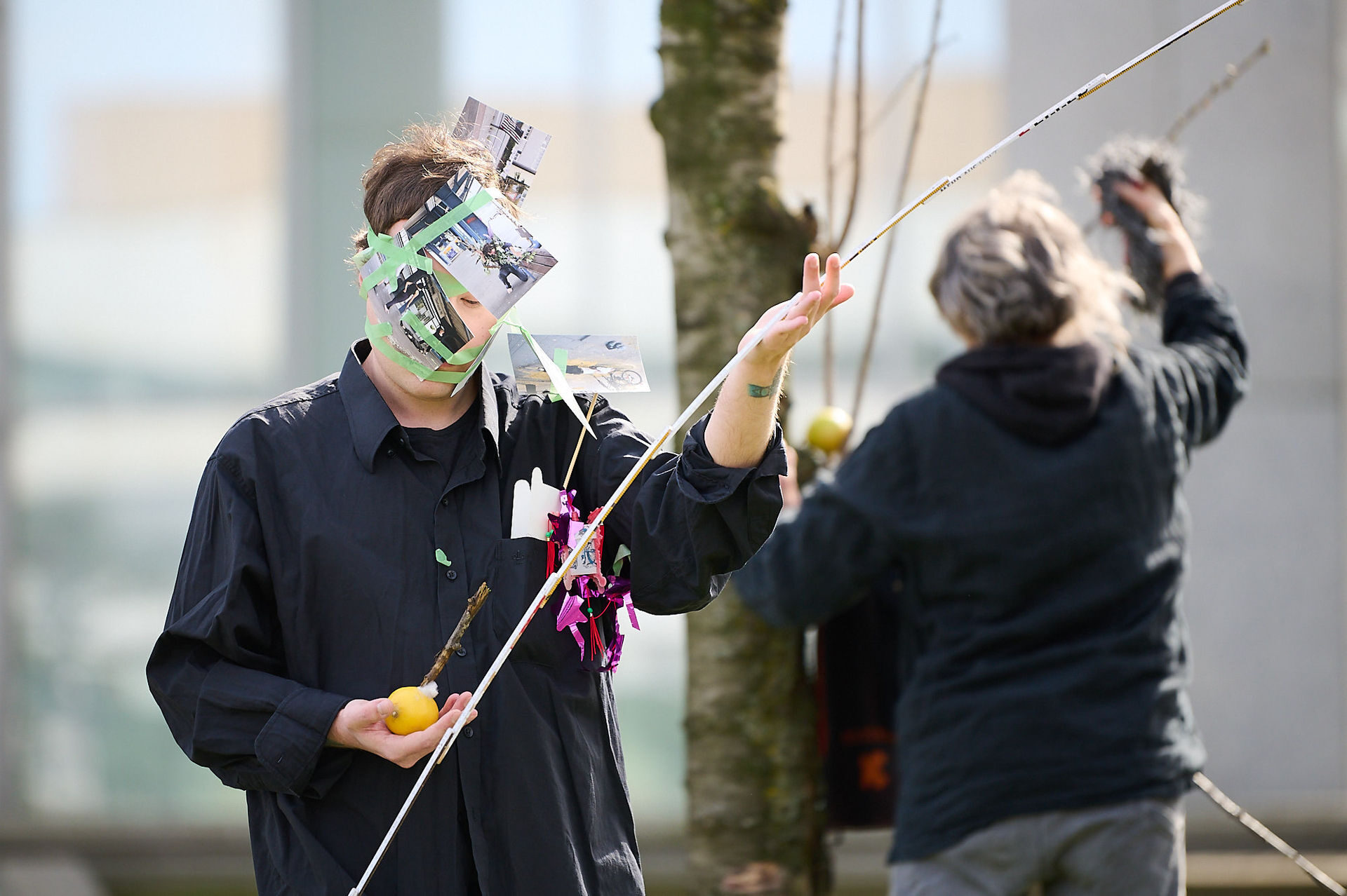 Two individuals engaged in an outdoor performance, one wearing a mask made of photos and holding a fruit, while the other interacts nearby.