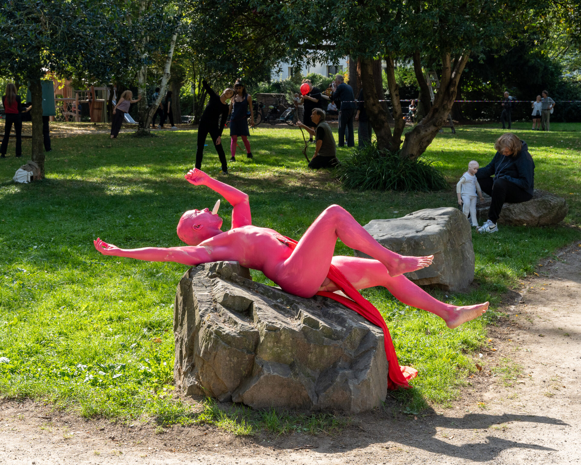 Eine Person mit leuchtend pinker Körperfarbe liegt auf einem Felsen in einem Park, umgeben von Grünflächen und Menschen, die verschiedenen Aktivitäten nachgehen.