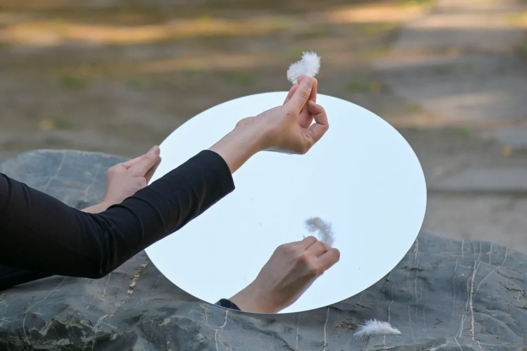 A hand holds delicate feathers above a round mirror, reflecting a serene outdoor setting with soft natural lighting.
