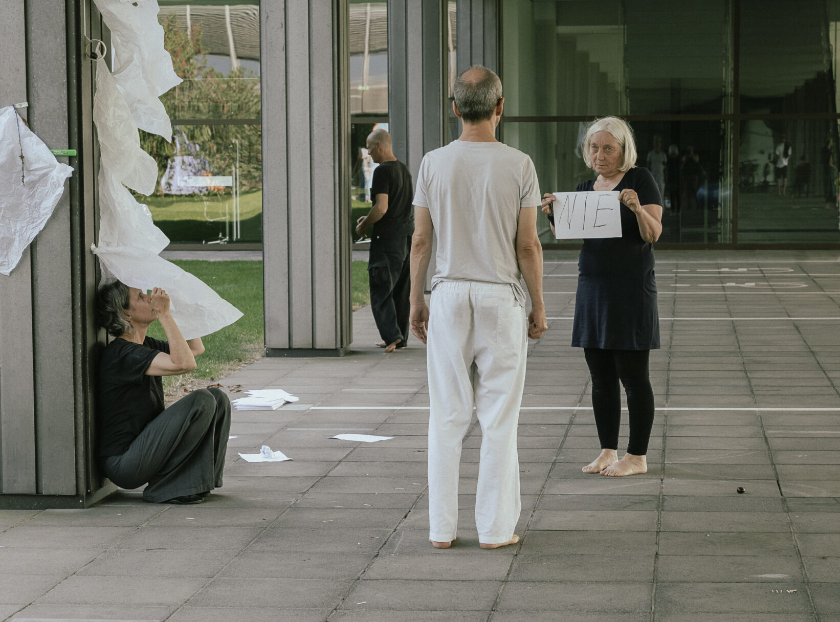 Three people in a modern outdoor space, one holding a sign, while others observe or interact, with scattered paper around.