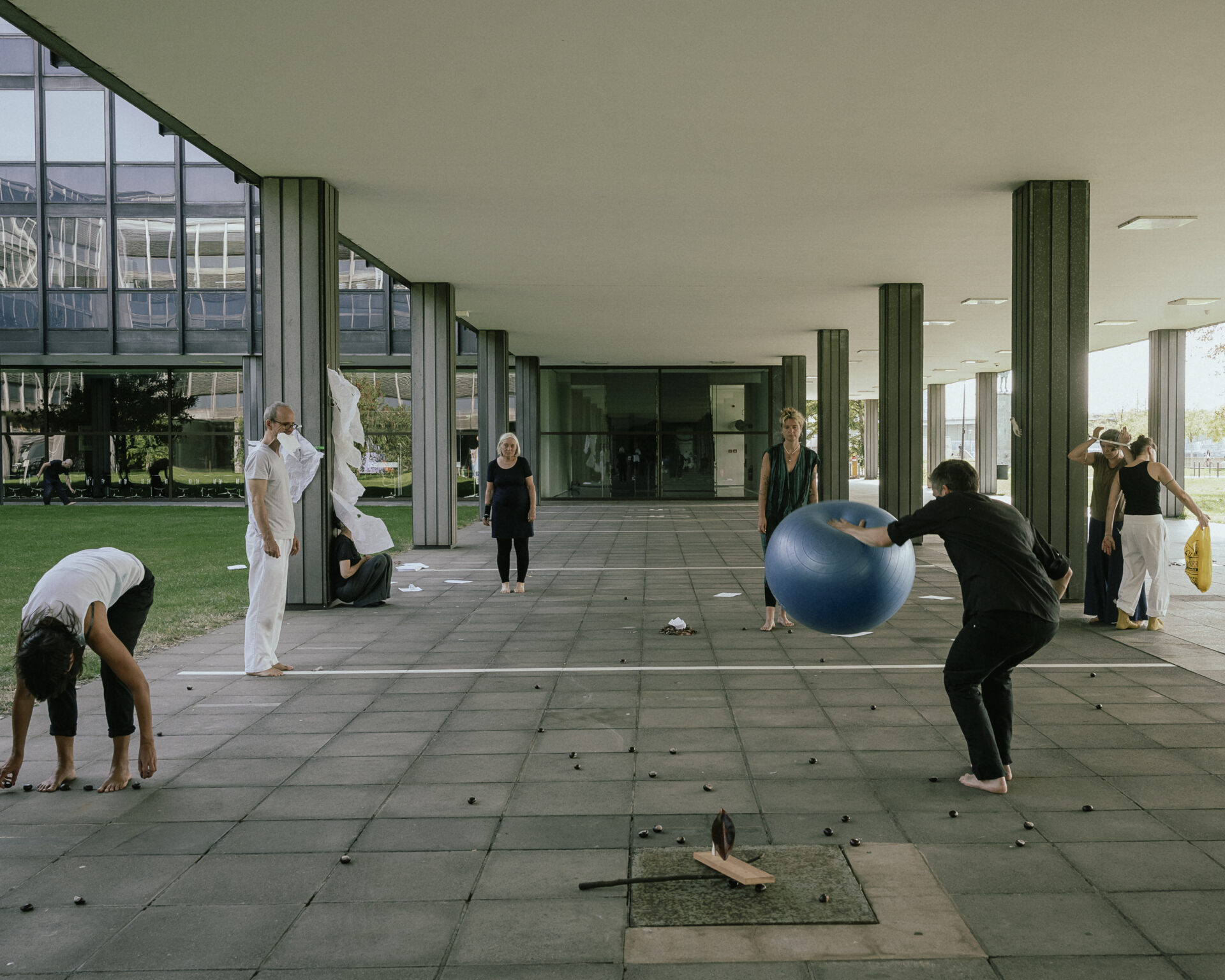Group of people engaging in playful activities outdoors, some stretching and others playing with a large blue ball, amid modern architecture.