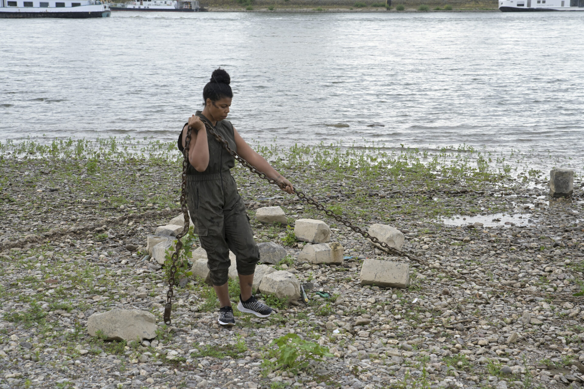 Person in green overalls stands on a rocky shoreline, pulling chains with water and boats in the background.