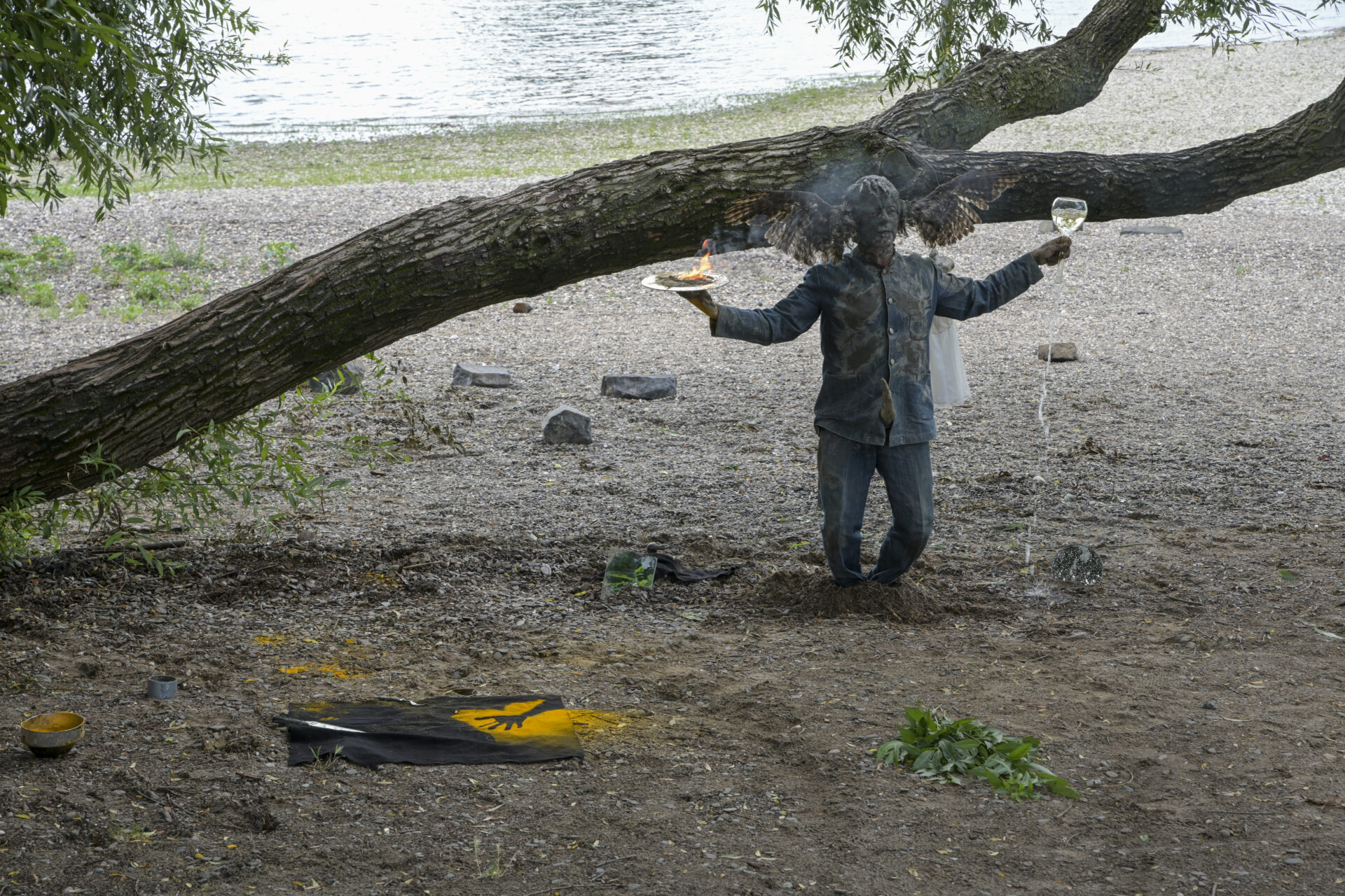 A figure in a raincoat stands in a shallow river, holding a glass in each hand, with a fallen tree overhead and scattered objects nearby.