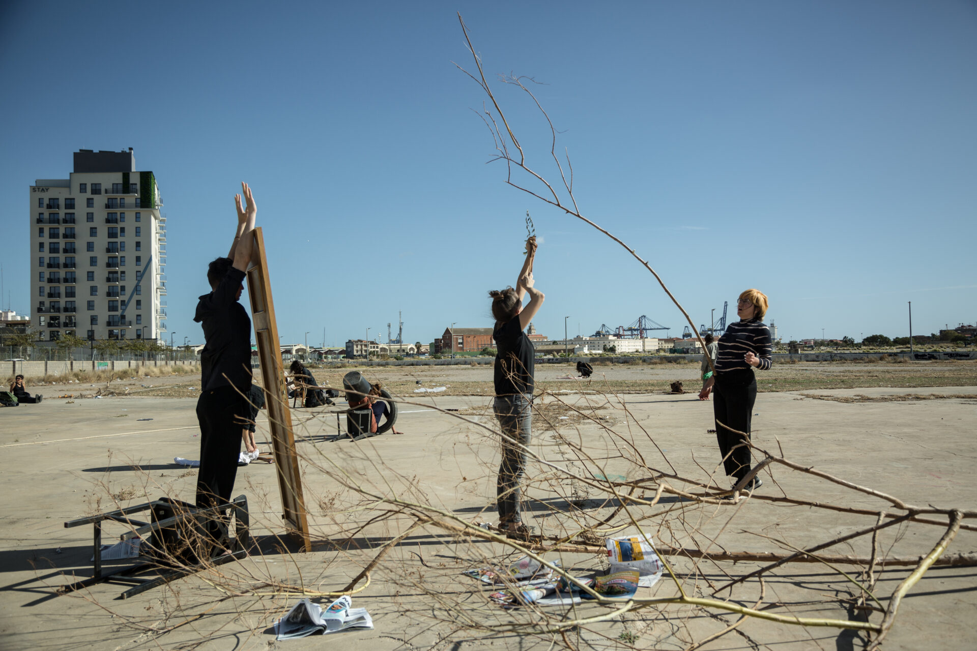 Three people work together on a sandy area, manipulating branches and objects, with a city skyline and clear blue sky in the background.