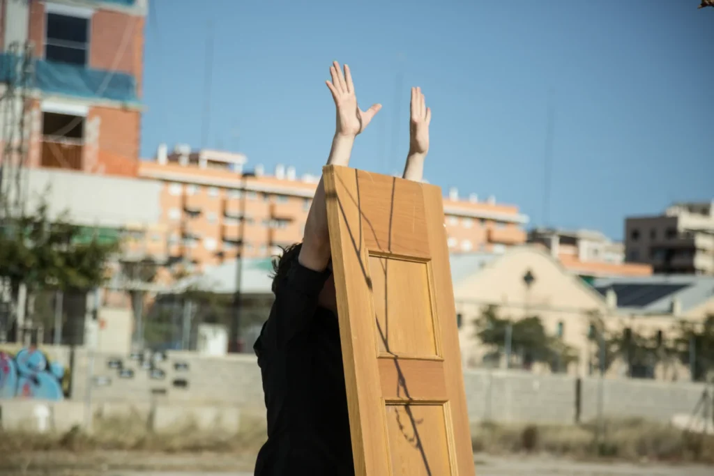 Person standing behind a wooden panel, raising hands towards the blue sky, in an urban outdoor setting.