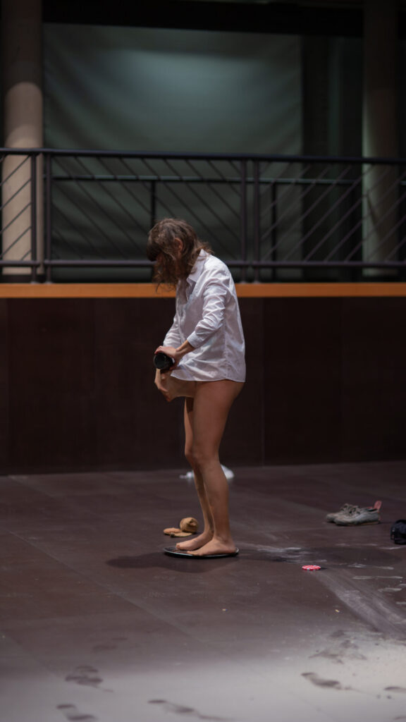 A person in a white shirt and flip-flops stands alone in a dimly lit space, looking down at their feet.