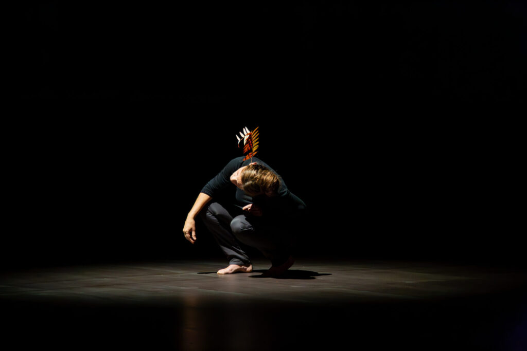 A person crouches in a spotlight on a dark stage, wearing a headpiece with feathers, conveying a sense of contemplation.