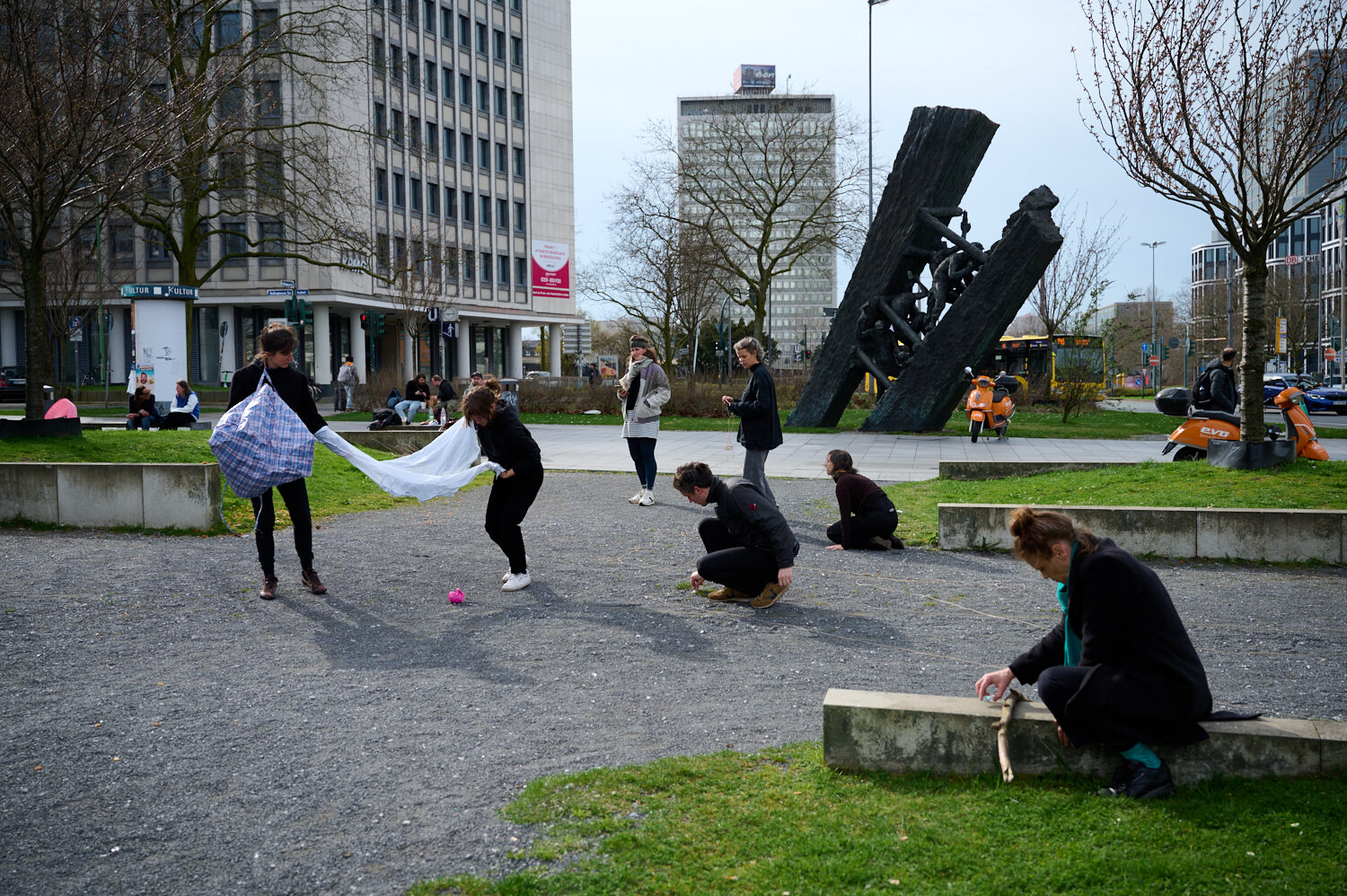 Group of people in a park engaging in a performance art project, with varied poses and expressions, surrounded by modern buildings.