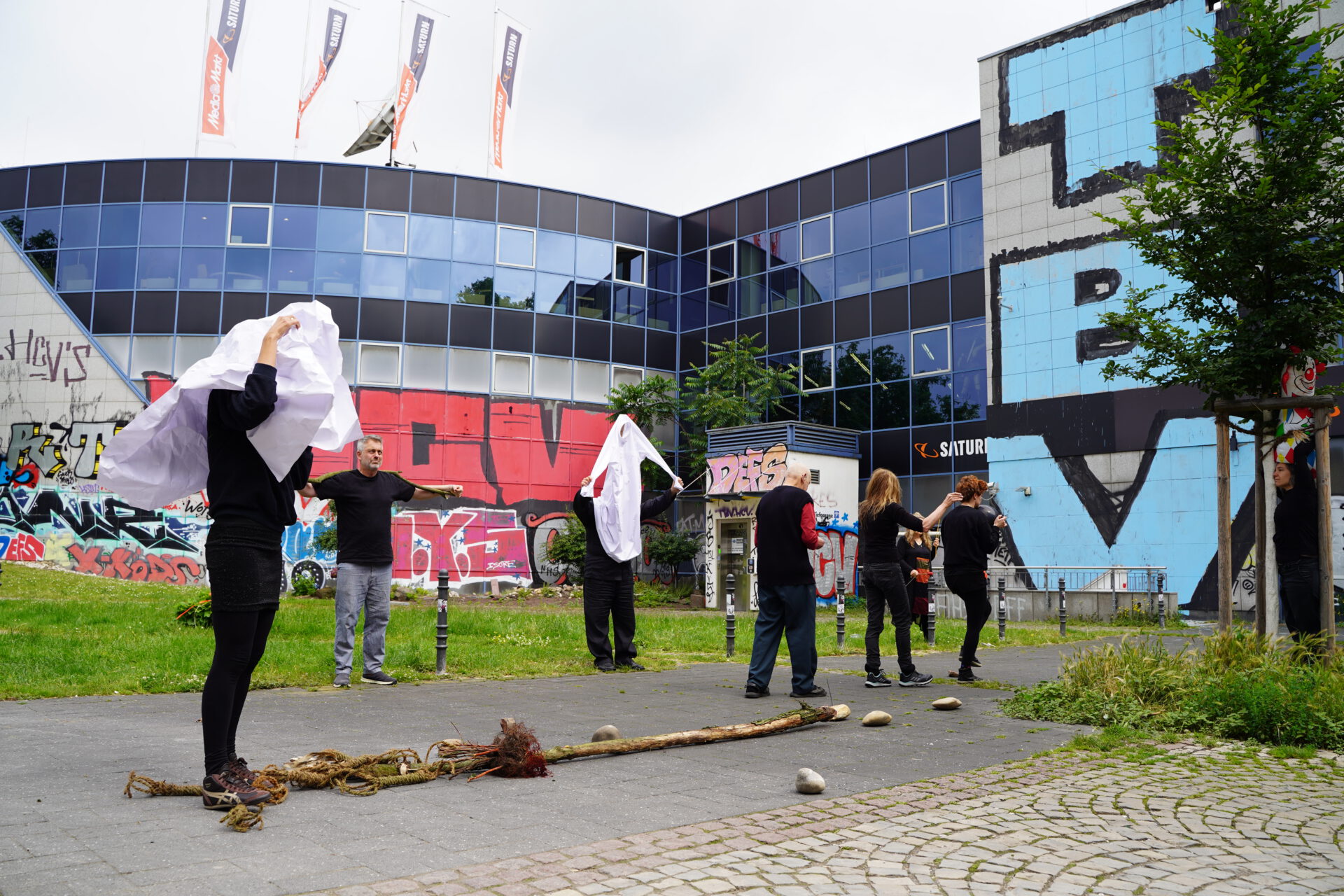 People gather outdoors near a graffiti-adorned building, some holding white cloths, while others arrange natural materials on the ground.