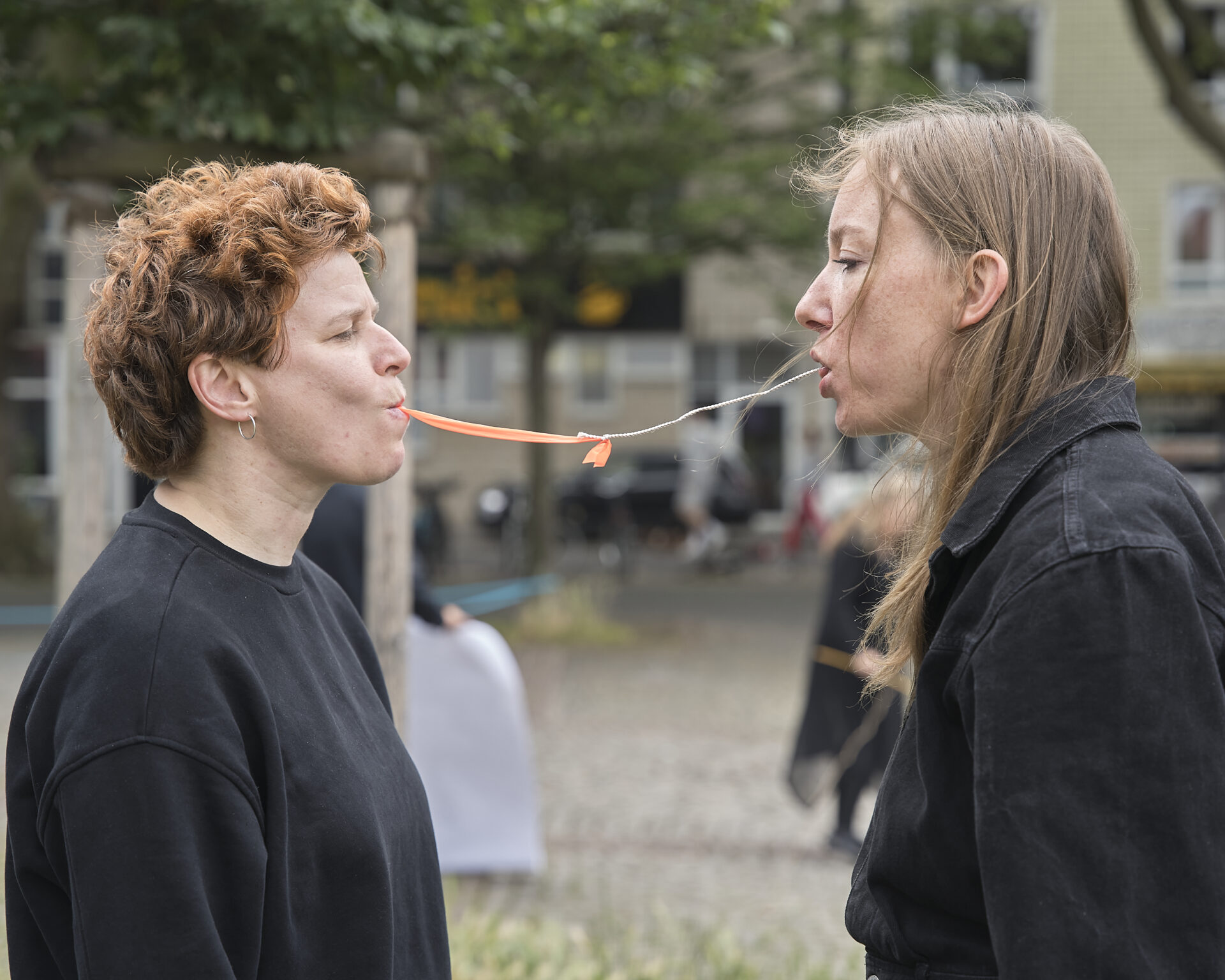 Two women face each other, each holding a piece of string in their mouths, with a playful expression amid a city setting.