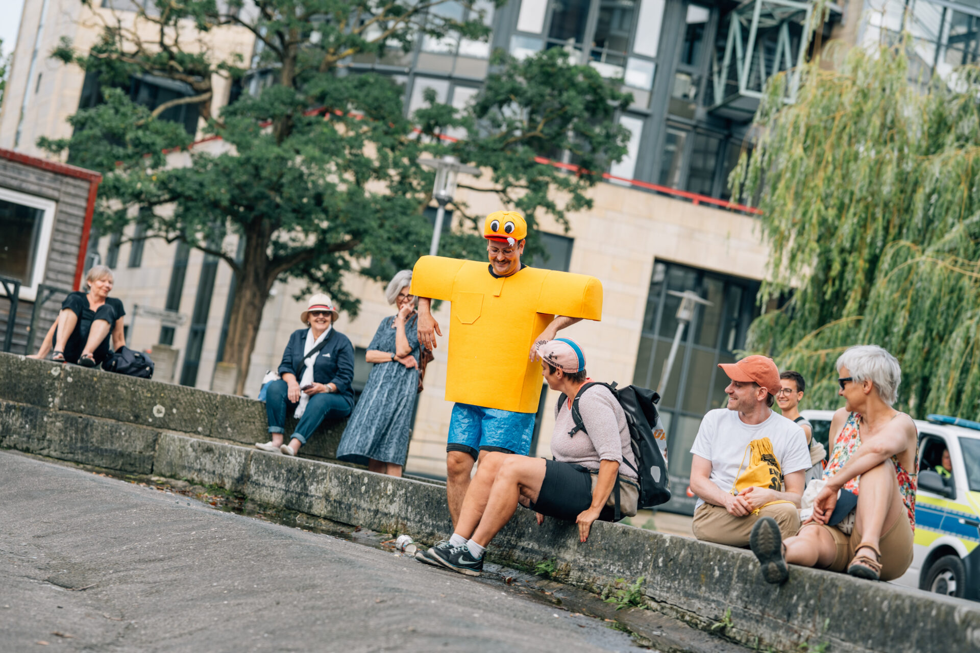 A person in a bright yellow inflatable costume poses playfully near a group of relaxed spectators in a lively urban setting.