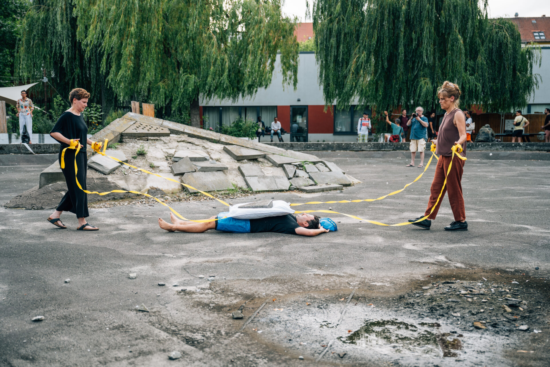 Two individuals in casual attire pull a third person lying on the ground with ropes, against a backdrop of a stone structure and spectators.