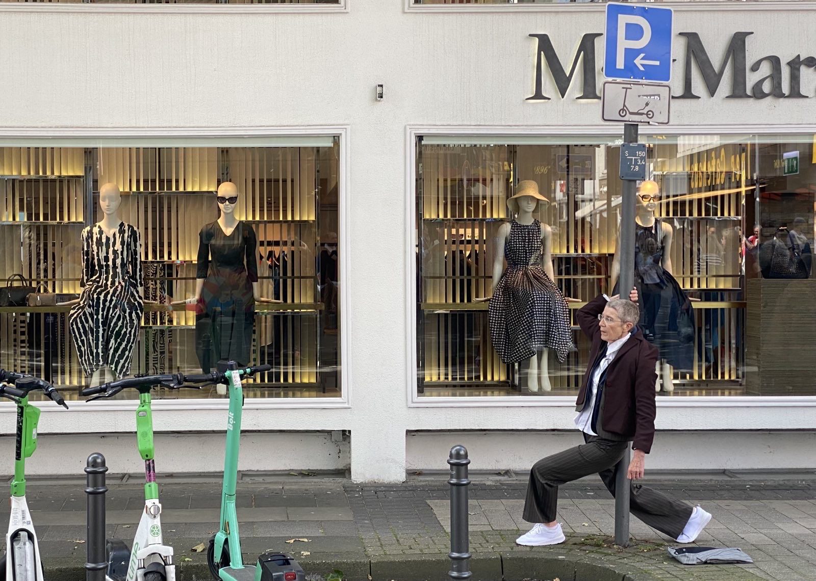 A woman in casual clothes stretches by a shop window displaying elegant dresses, with scooters parked nearby.