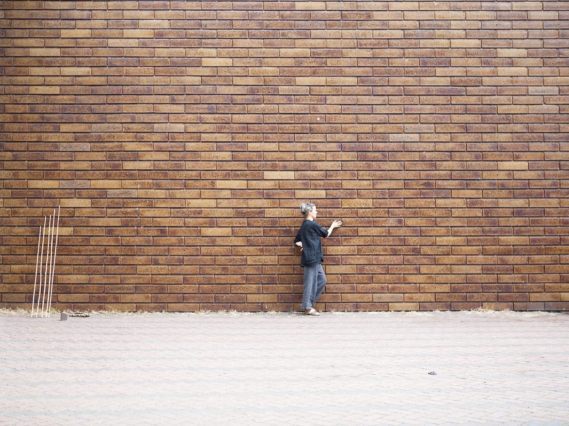 Child in casual clothes stands against a brown brick wall, appearing contemplative, with wooden sticks on the ground nearby.