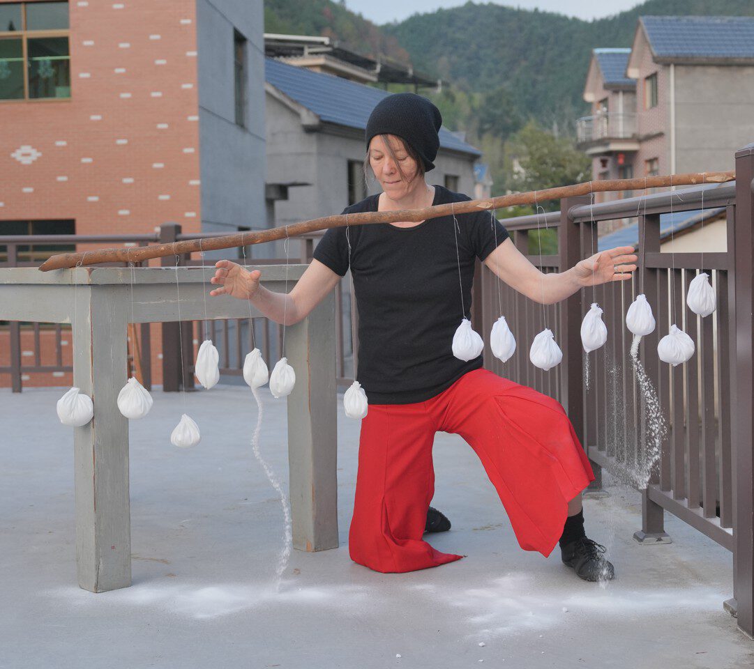 A person in a black shirt and red pants balances on one knee, manipulating hanging white objects on a stick in an outdoor setting.