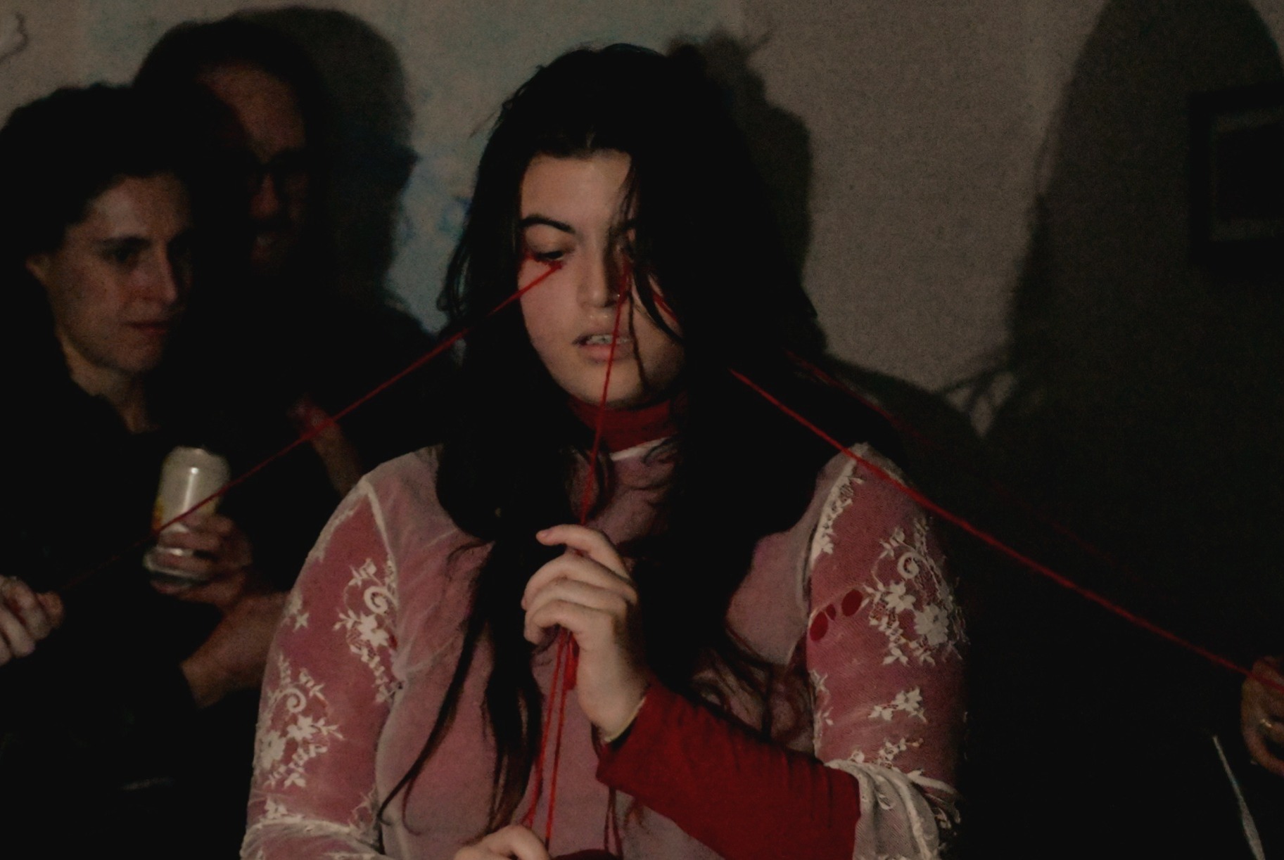 Person with long black hair with red string in their hands that eminates from their eyes outwards and held by members of the audience